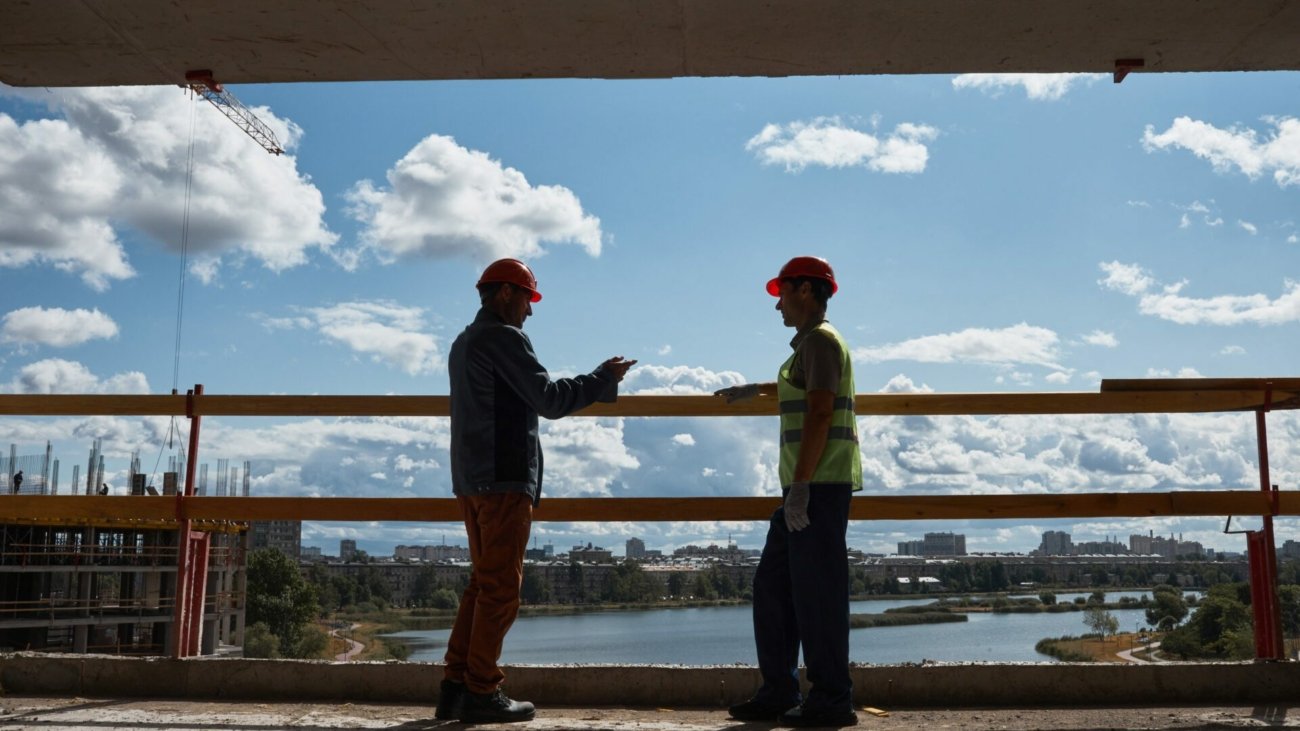 Backlit side view at two construction workers discussing project while standing at high rise building, copy space
